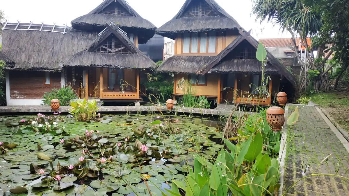 Bungalow beratap ijuk berdiri di seberang kolam teratai yang sedang berbunga